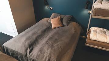 Una cama individual con una colcha texturizada y una almohada, una lámpara de noche y un estante en la pared con mantas dobladas.