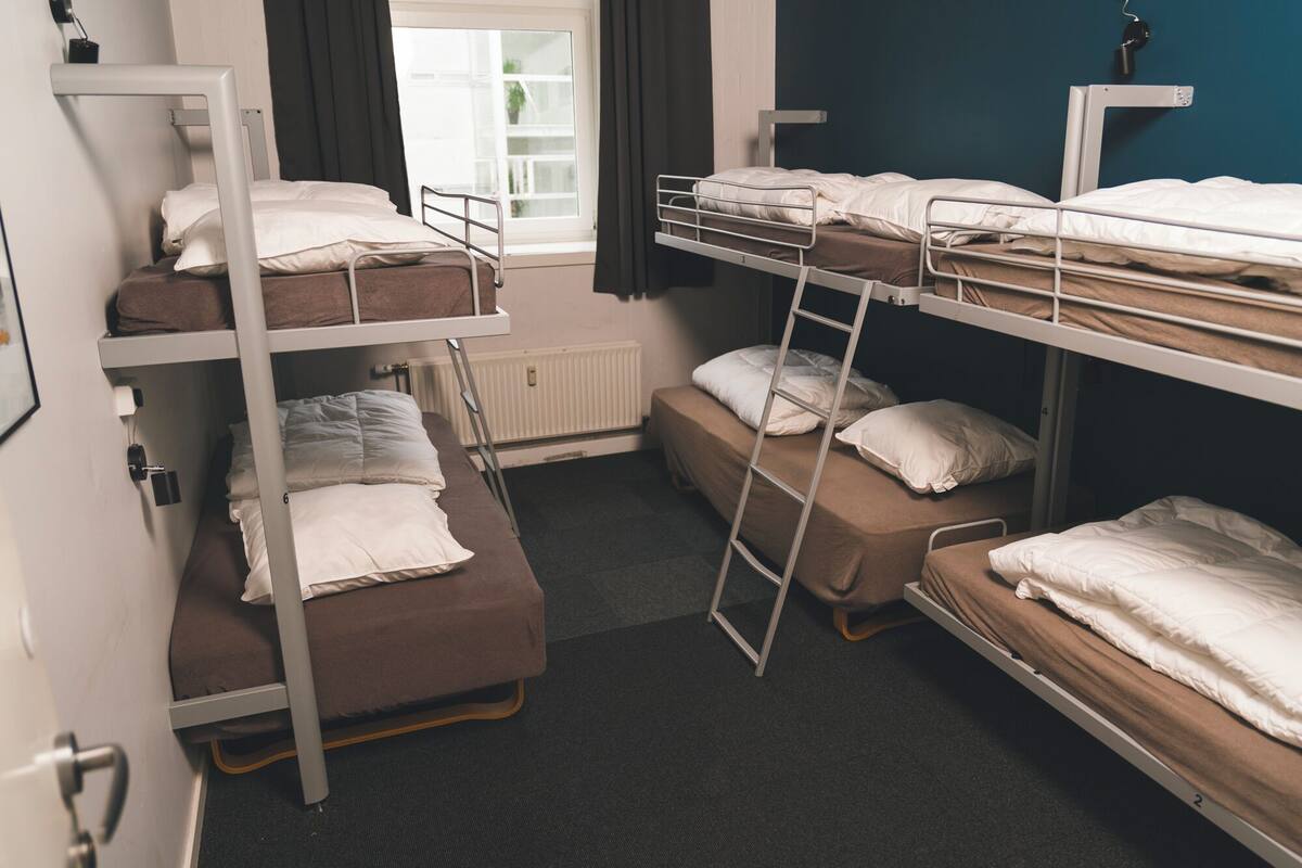 A dormitory room with bunk beds, a radiator, a window with curtains, and a door.