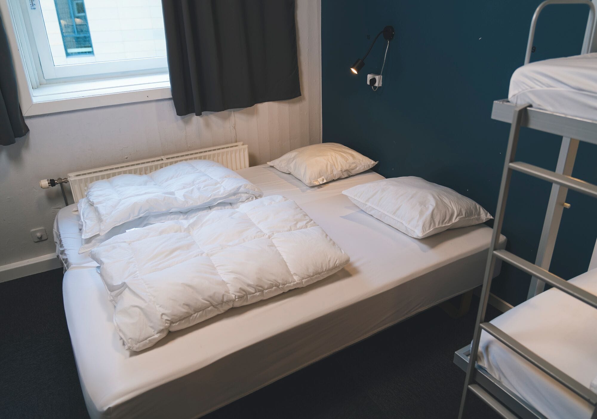 A single bed with white bedding, a window with curtains, a radiator, and a wall-mounted lamp.