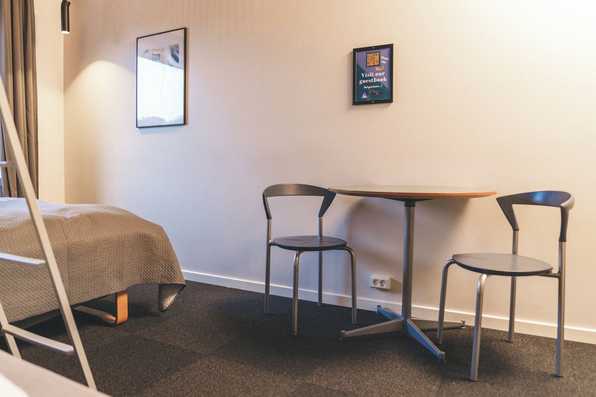 Una habitación pequeña con una cama, una mesa y dos sillas. Hay un cuadro en la pared y un letrero con un código QR.