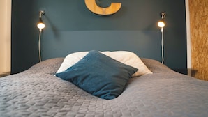 Una cama con un cubrecolchón gris acolchado, dos almohadas y una gran 'C' de madera en la pared.