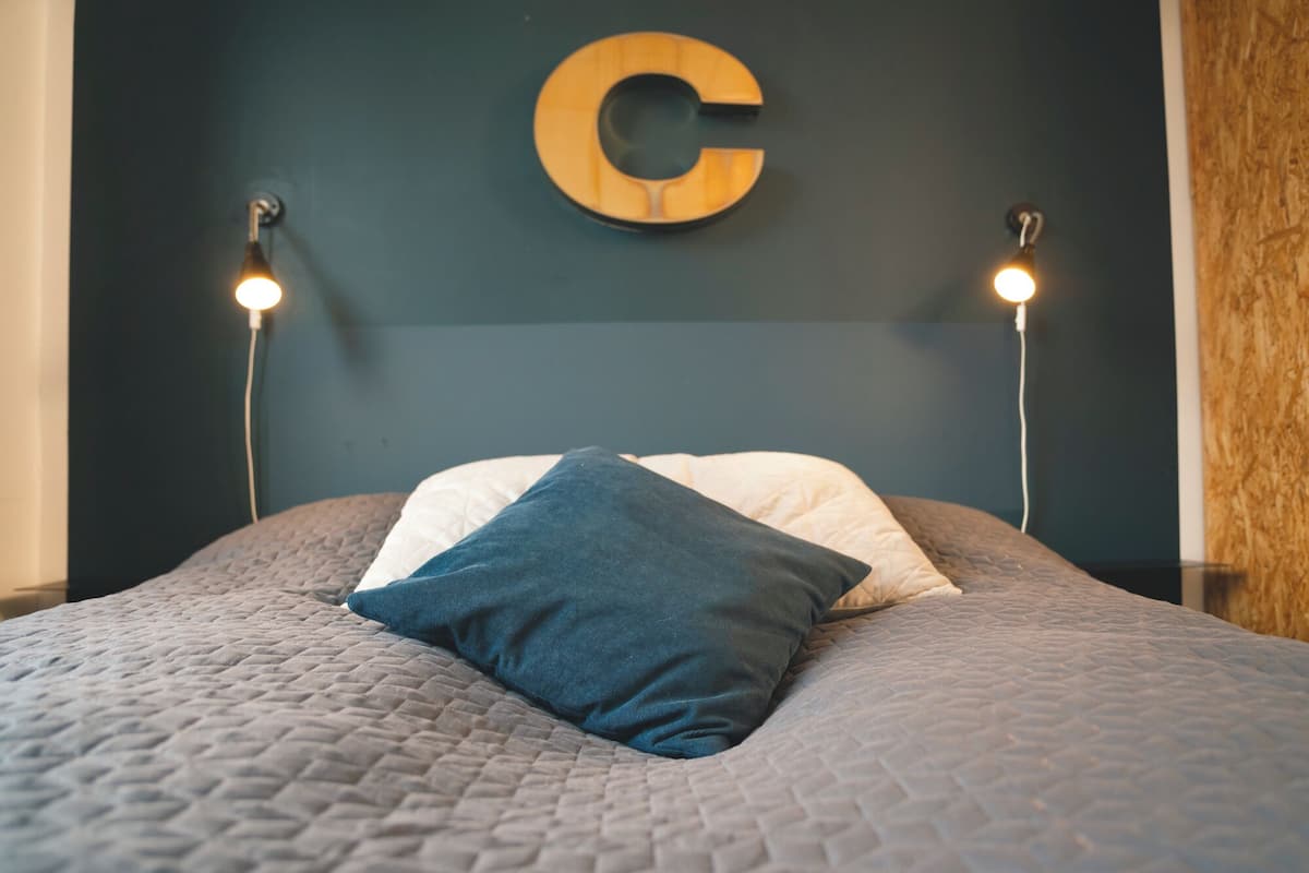 A bed with a quilted gray bedspread, two pillows, and a large wooden 'C' on the wall.