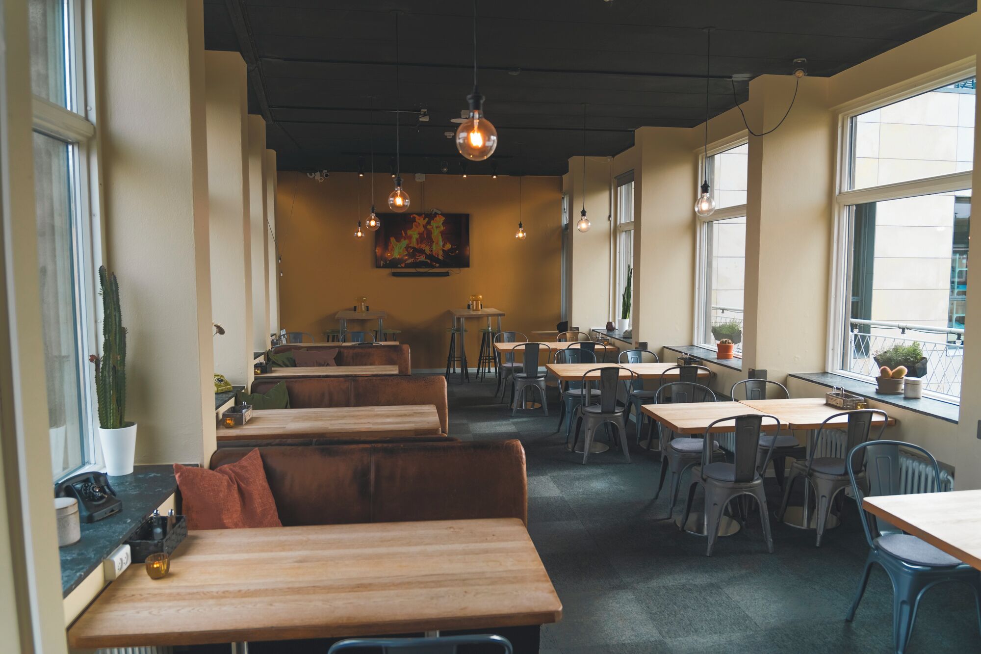 A restaurant interior with wooden tables and benches, large windows, and hanging pendant lights.