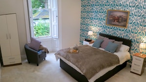 A bedroom with a large bed, bedside tables, a chair, a wardrobe, and a window with a view of greenery. - Inverleith Estate (Edinburgh)