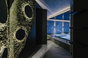 A modern bathroom with a large, textured wall, a visible bathtub, and a glass partition. - Hotel Freina (Selva di Val Gardena)