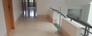 A hallway with a wooden door on the left, a metal railing on the right, and a staircase leading to an upper floor. - Hotel Hemala (Karur)