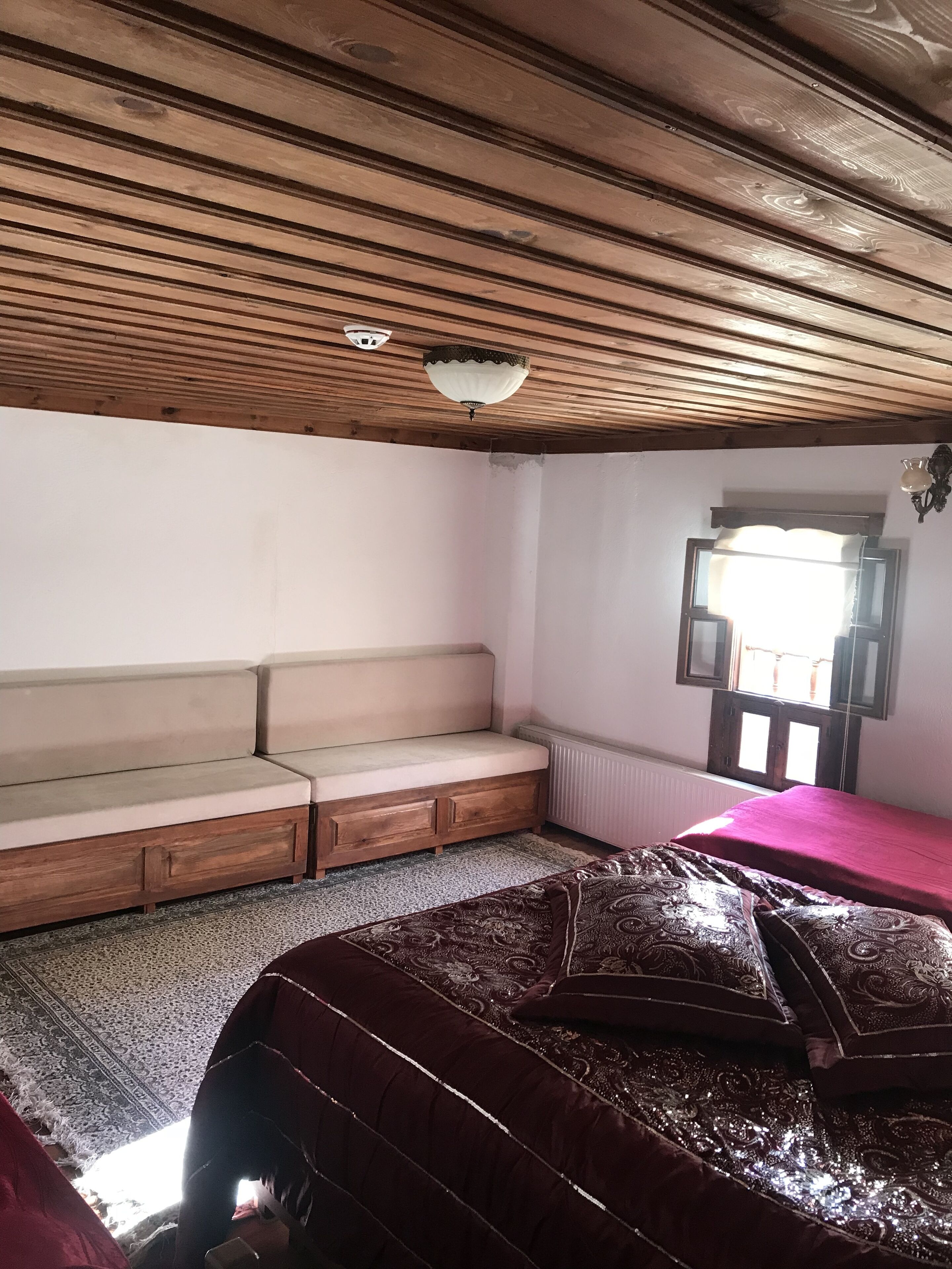 Een kamer met een houten plafond, een bank, een bed met beddengoed met een patroon, een raam met gordijnen, en een radiator.