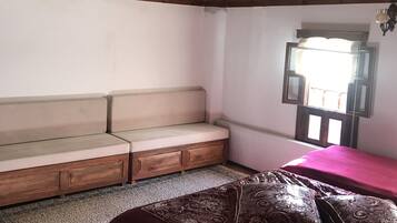 Een kamer met een houten plafond, een bank, een bed met beddengoed met een patroon, een raam met gordijnen, en een radiator.