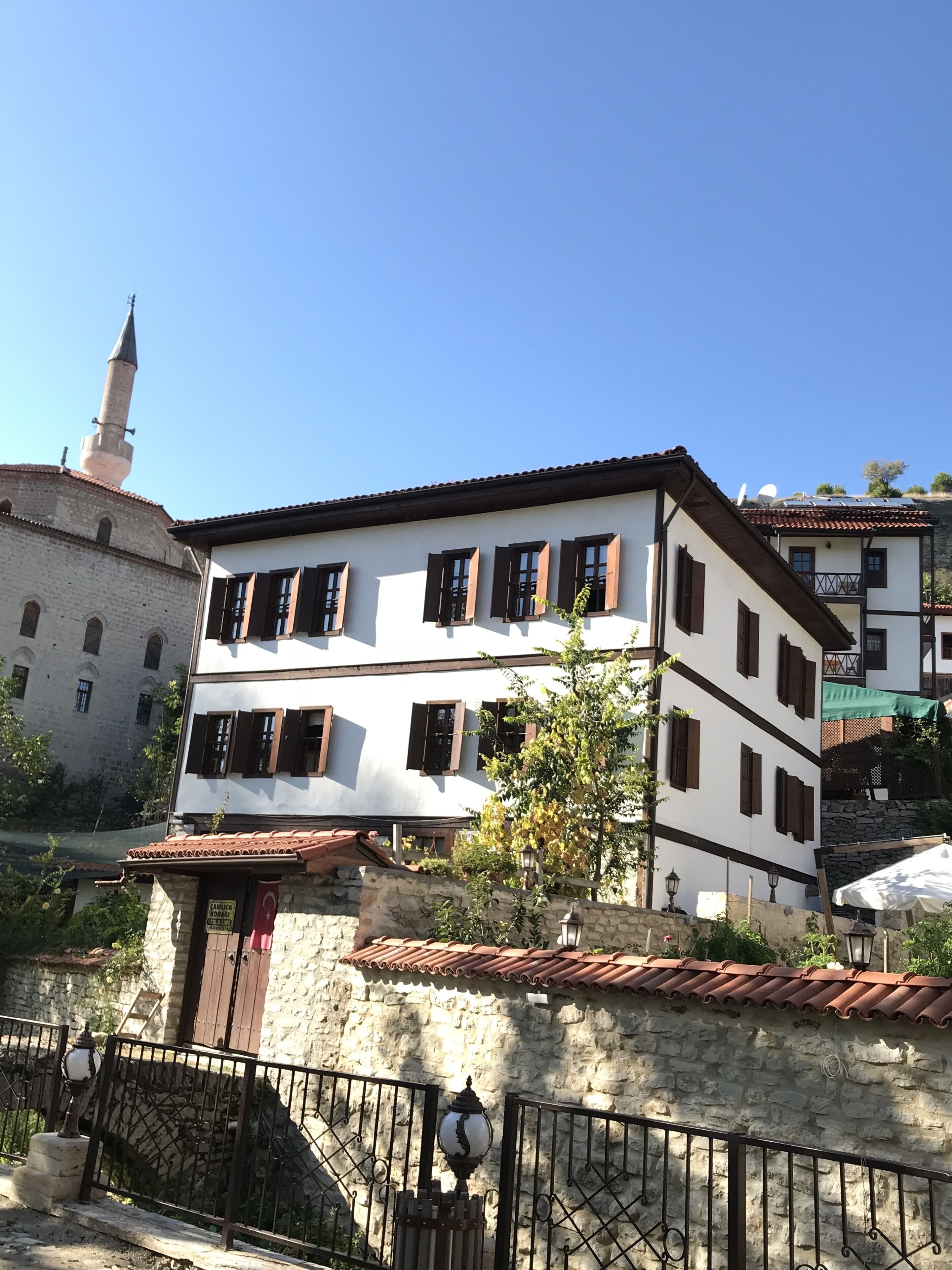 A two-story building with a white facade and brown shutters, a stone wall with a gate, and a mosque with a minaret in the background.