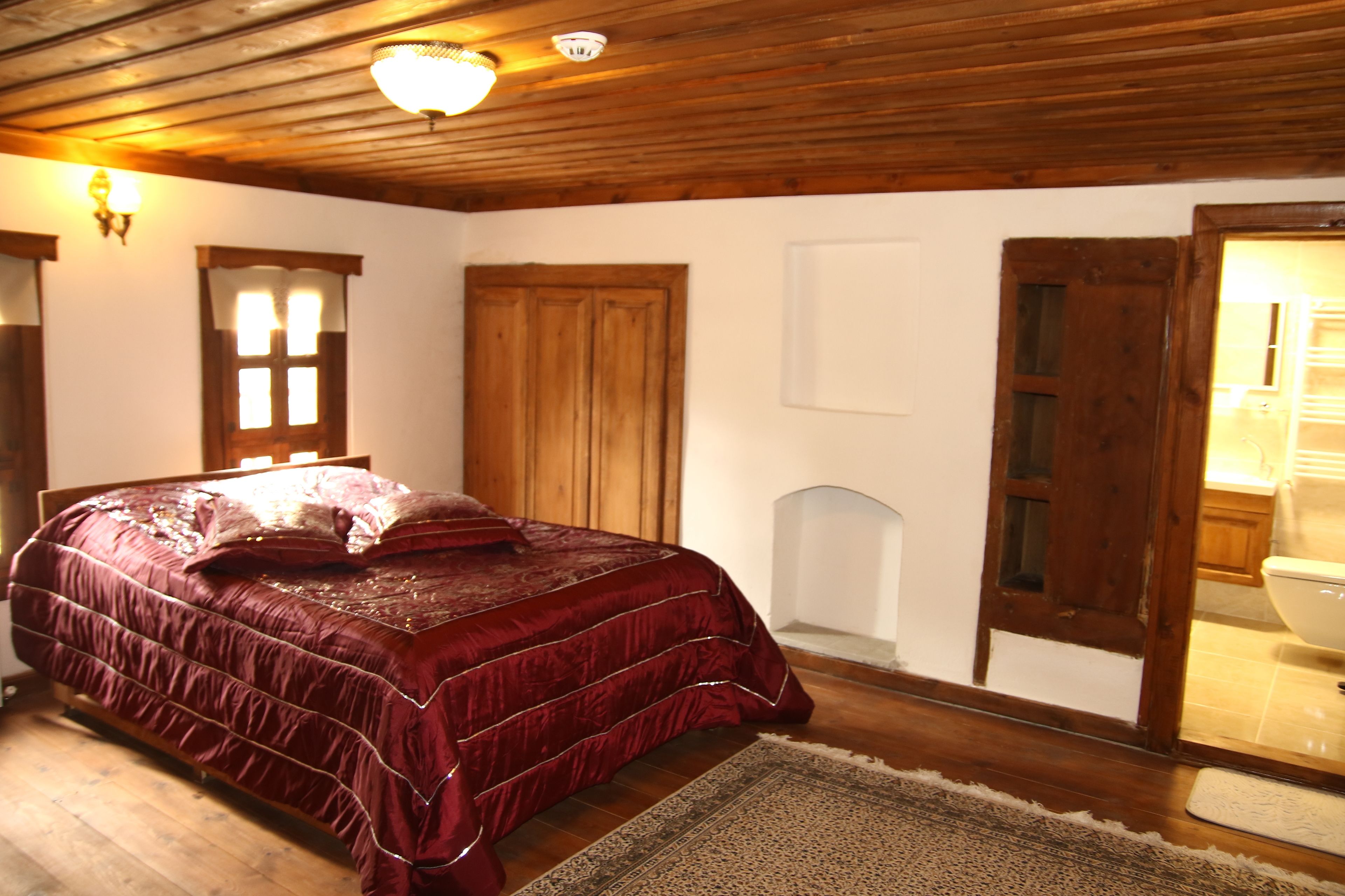 A bedroom with a wooden ceiling, a large bed with a red bedspread, a wooden wardrobe, and a white door leading to a bathroom.
