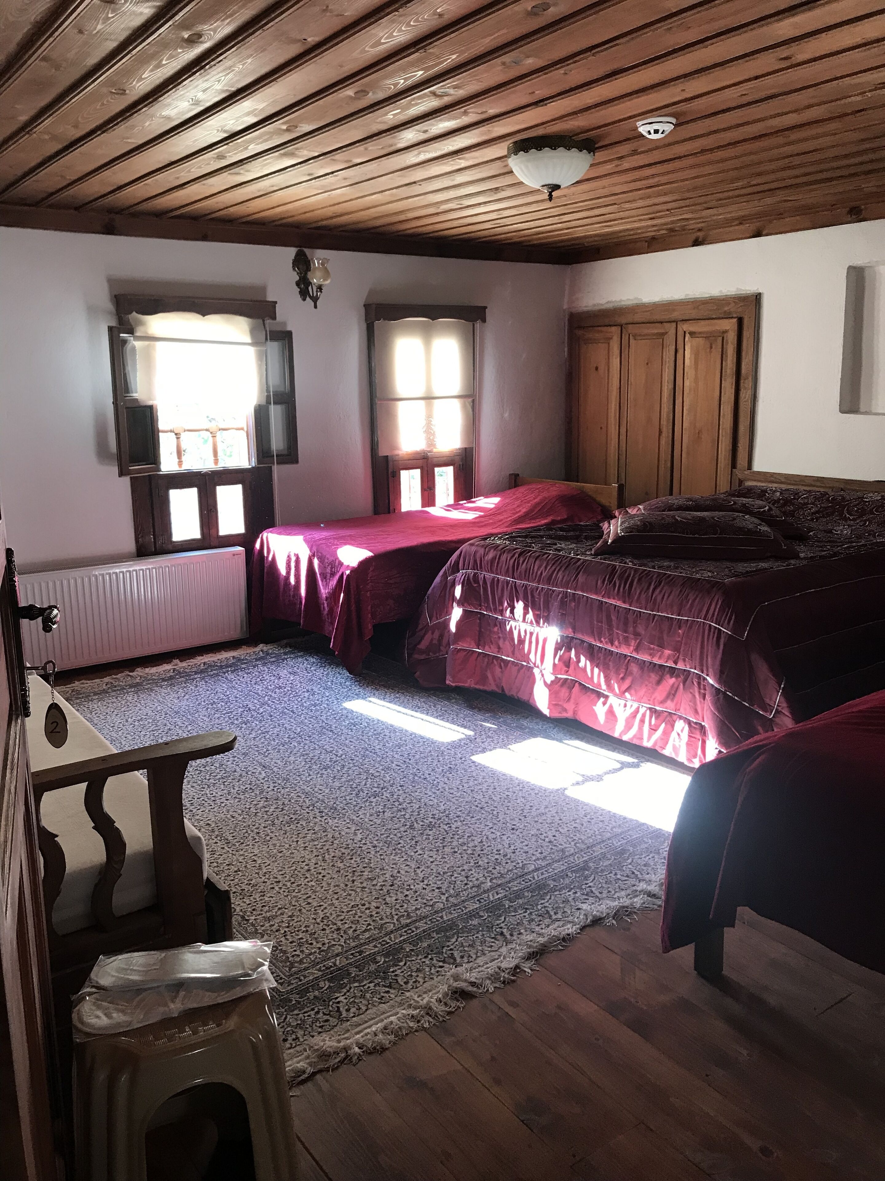 A room with wooden ceiling beams, a bed with a red bedspread, a wooden wardrobe, and a window with a white blind.