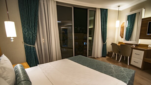 A hotel room with a bed, a desk with a chair, a TV, and a sliding glass door leading to a balcony. - Exelans Hotel (Fethiye)