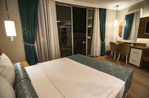 A hotel room with a bed, a desk with a chair, a TV, and a sliding glass door leading to a balcony. - Exelans Hotel (Fethiye)
