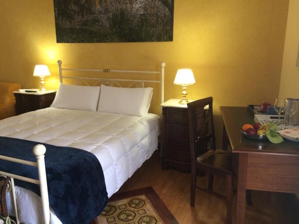 A hotel room with a bed, two bedside lamps, a wooden nightstand, a chair, a small table with a fruit bowl, and a painting on the wall.
