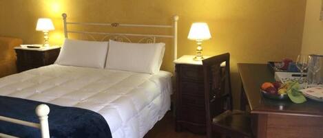 A hotel room with a bed, two bedside lamps, a wooden nightstand, a chair, a small table with a fruit bowl, and a painting on the wall.