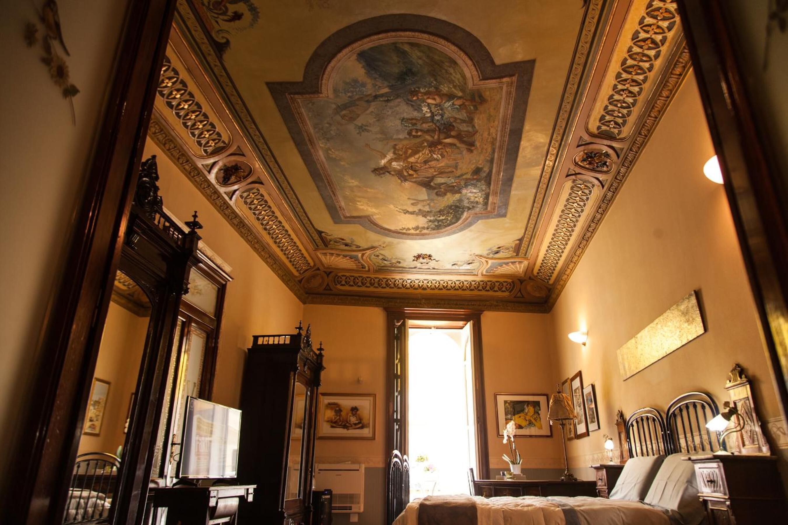A room with ornate ceiling frescoes, a bed, a desk with a computer, and a window allowing natural light.
