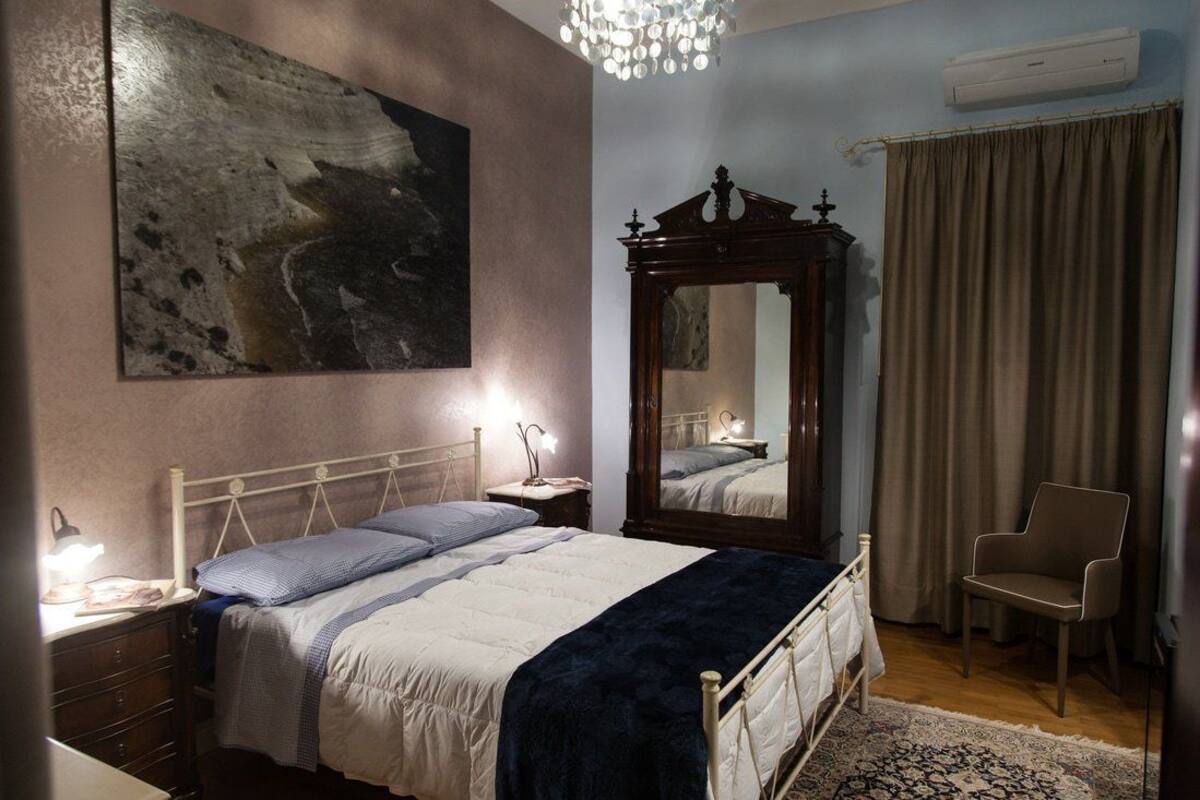 A bedroom with a bed, a nightstand, a dresser, a chair, a large mirror, a chandelier, and a painting on the wall.