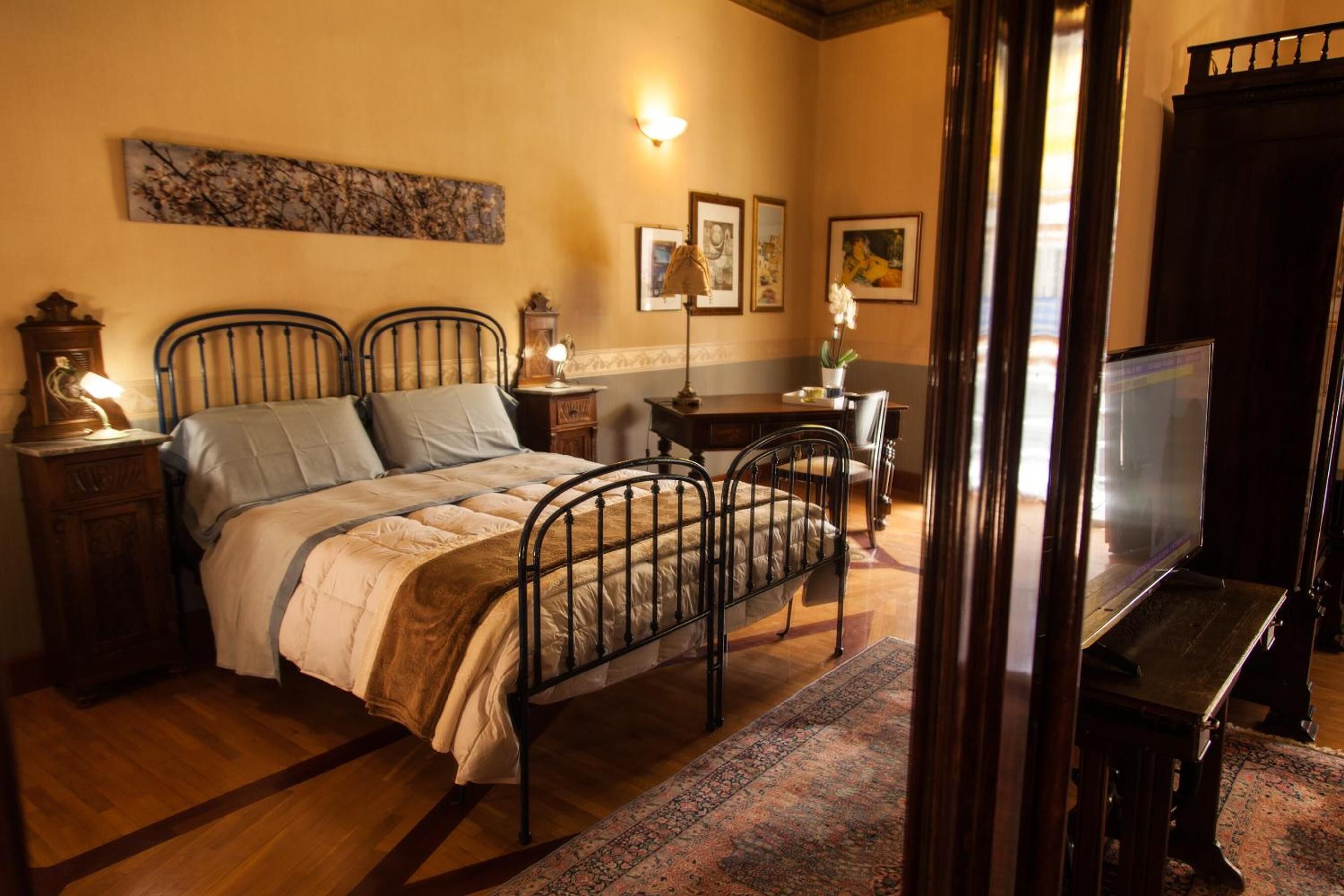 A bedroom with a bed, bedside tables, a desk, a chair, a television, and artwork on the walls.
