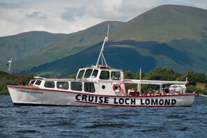 Imbarcazione chiamata 'Cruise Loch Lomond' con passeggeri a bordo, che naviga in acqua con montagne sullo sfondo.