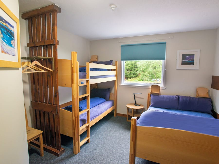Ein Schlafsaalzimmer mit Etagenbetten, einem kleinen Tisch, einem Fenster mit Blick ins Grüne und einem gerahmten Bild an der Wand.