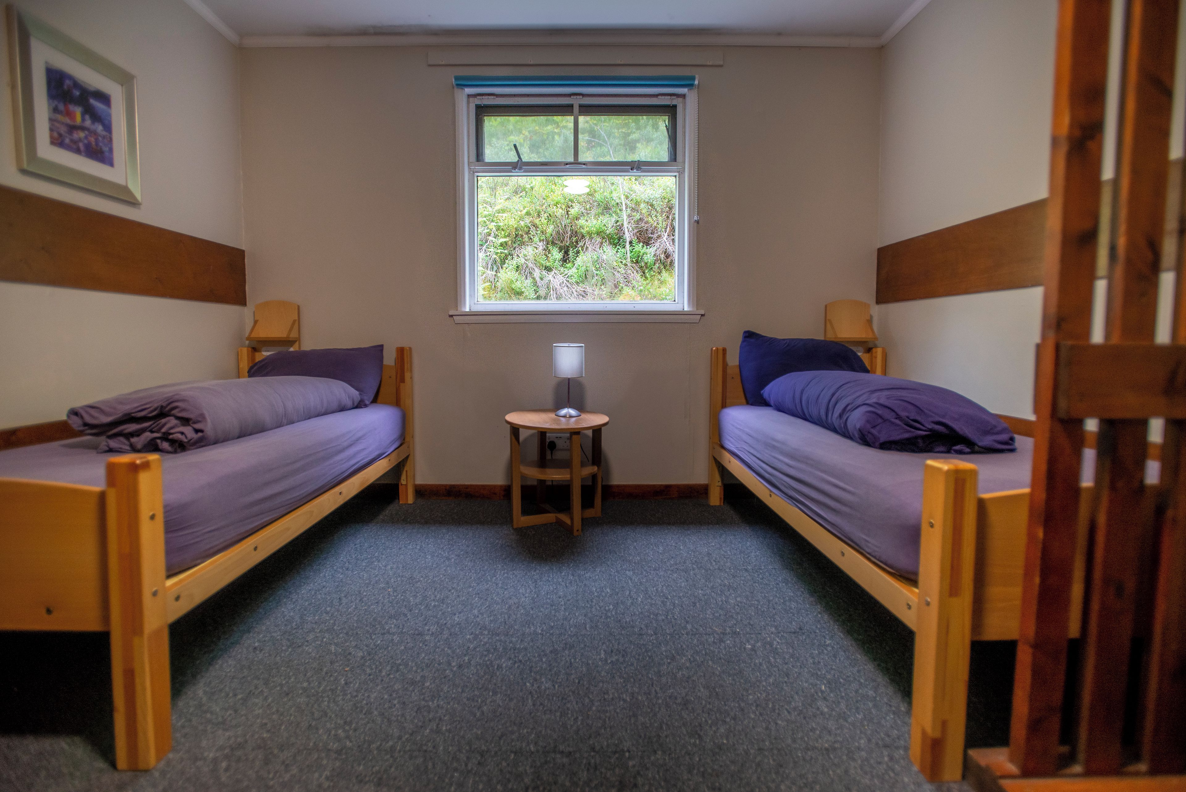 A small room with two wooden bunk beds, a small table with a lamp, and a window with a view of greenery.