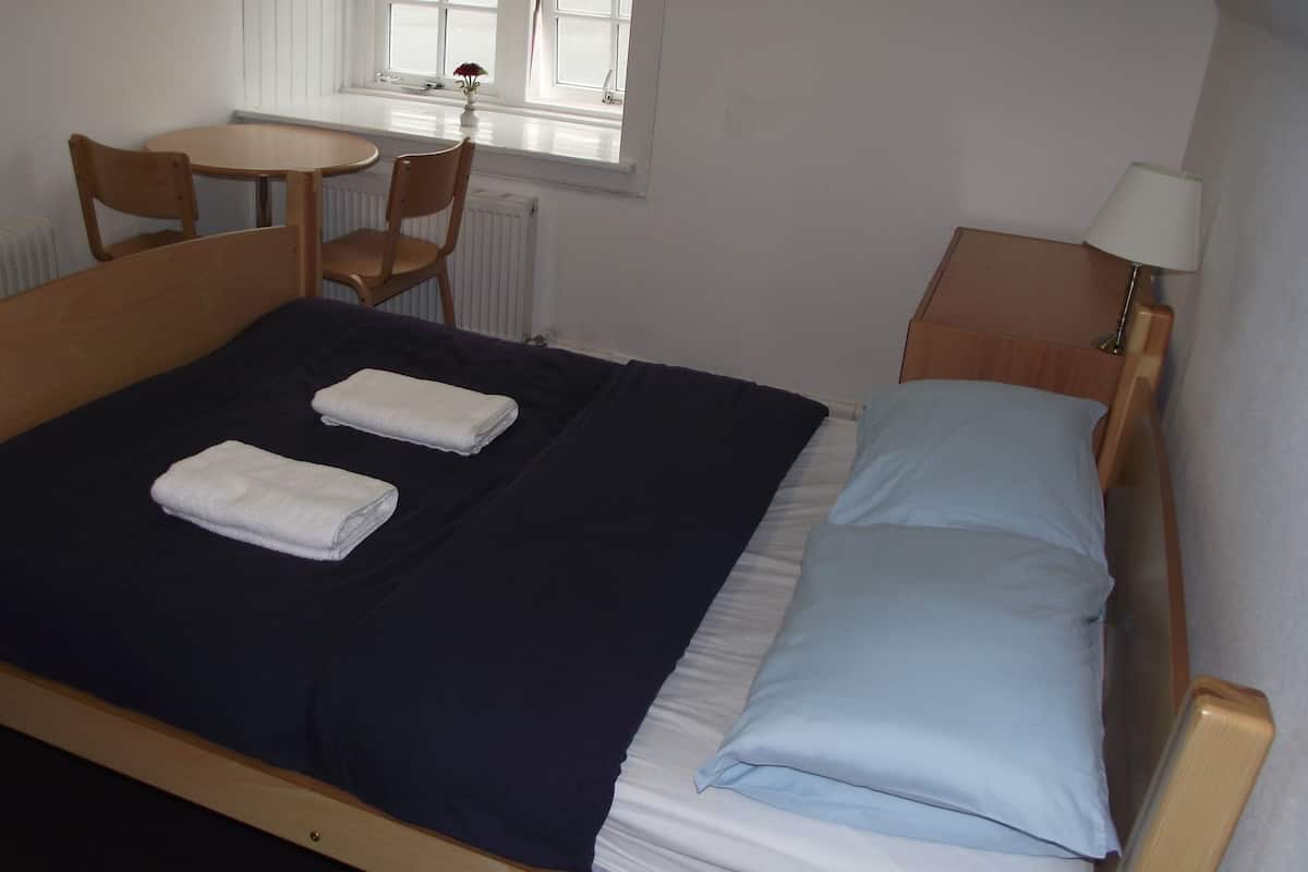 Ein Einzelbett mit zwei ordentlich gefalteten Handtüchern, ein kleiner runder Tisch mit einem Stuhl, eine Lampe und ein Fenster mit Blick.