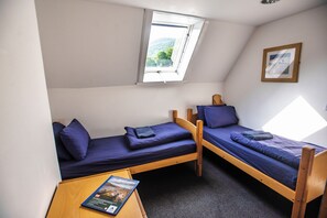 A small, compact room with two single beds, a window, a framed picture on the wall, and a table with a book. - Lochranza Youth Hostel (Isle of Arran)