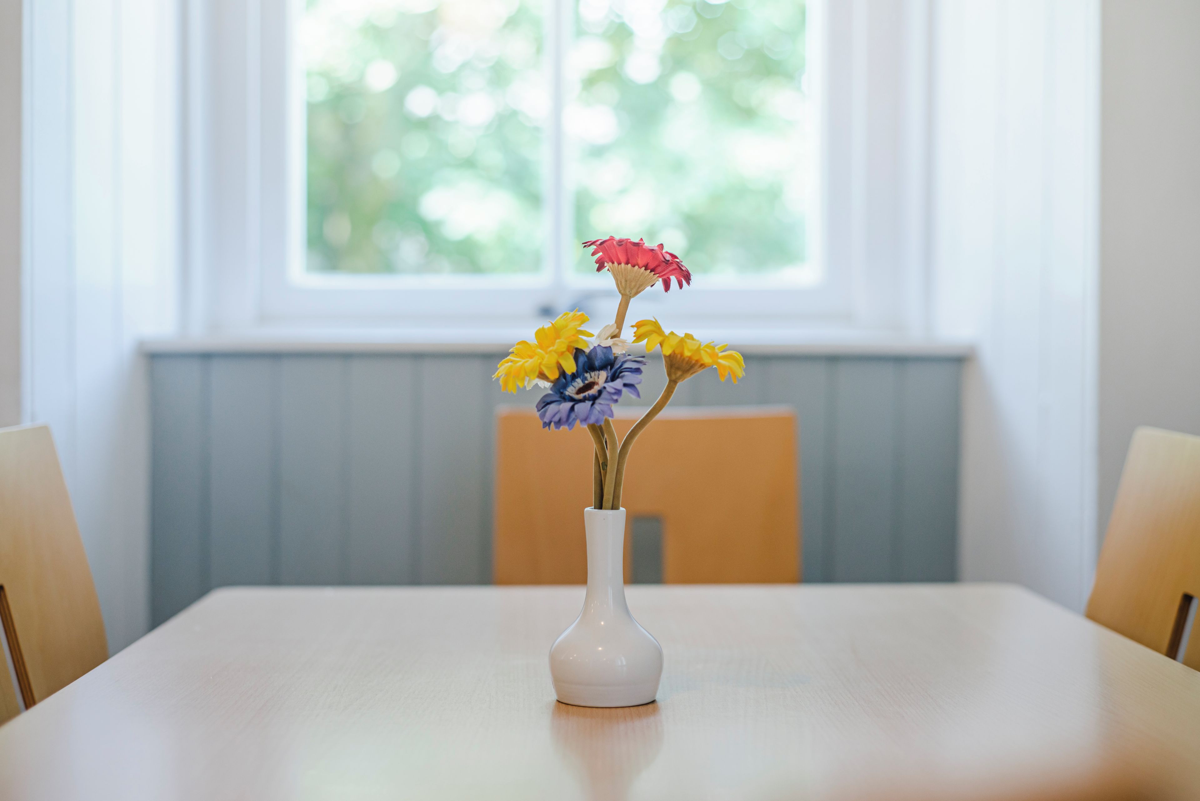 A white vase with flowers on a table in a room with a window.