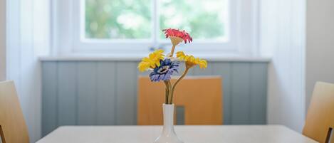 A white vase with flowers on a table in a room with a window.
