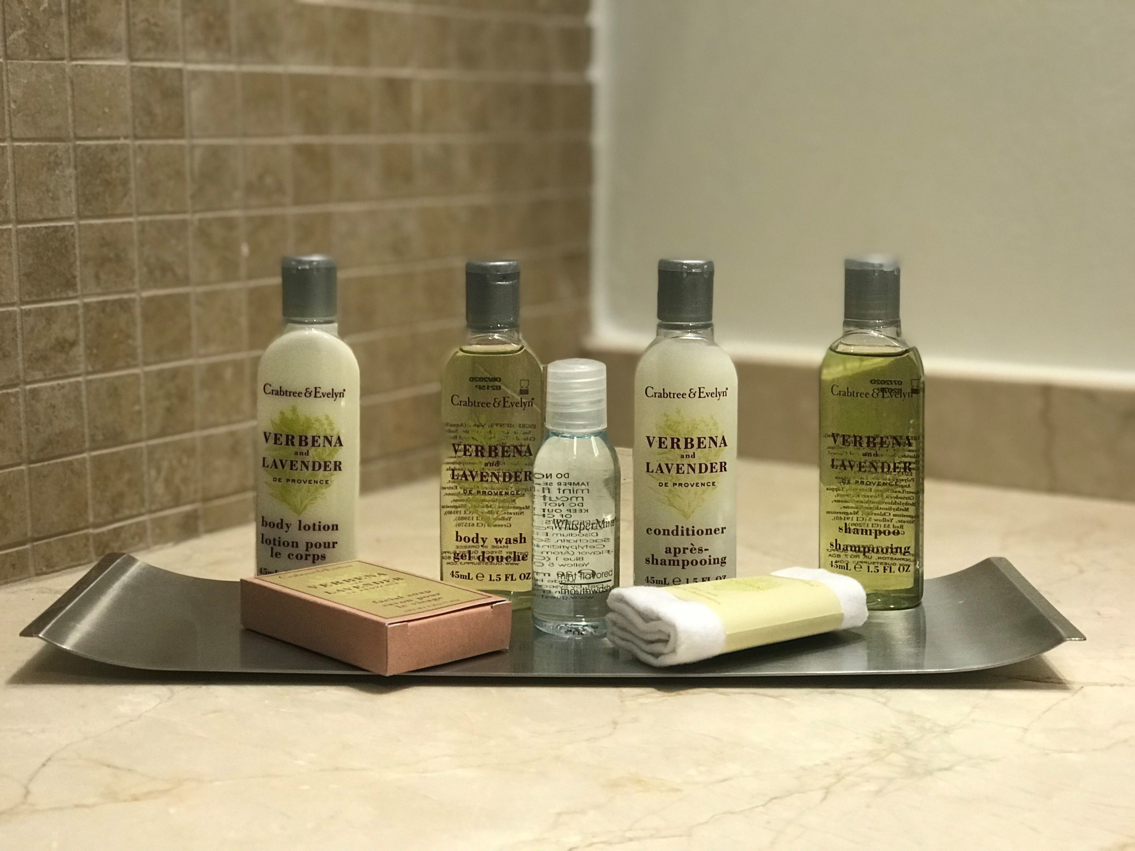 Una encimera de baño con cinco botellas de productos de cuidado corporal Verbenal de Crabtree & Evelyn, una pastilla de jabón y una toalla.