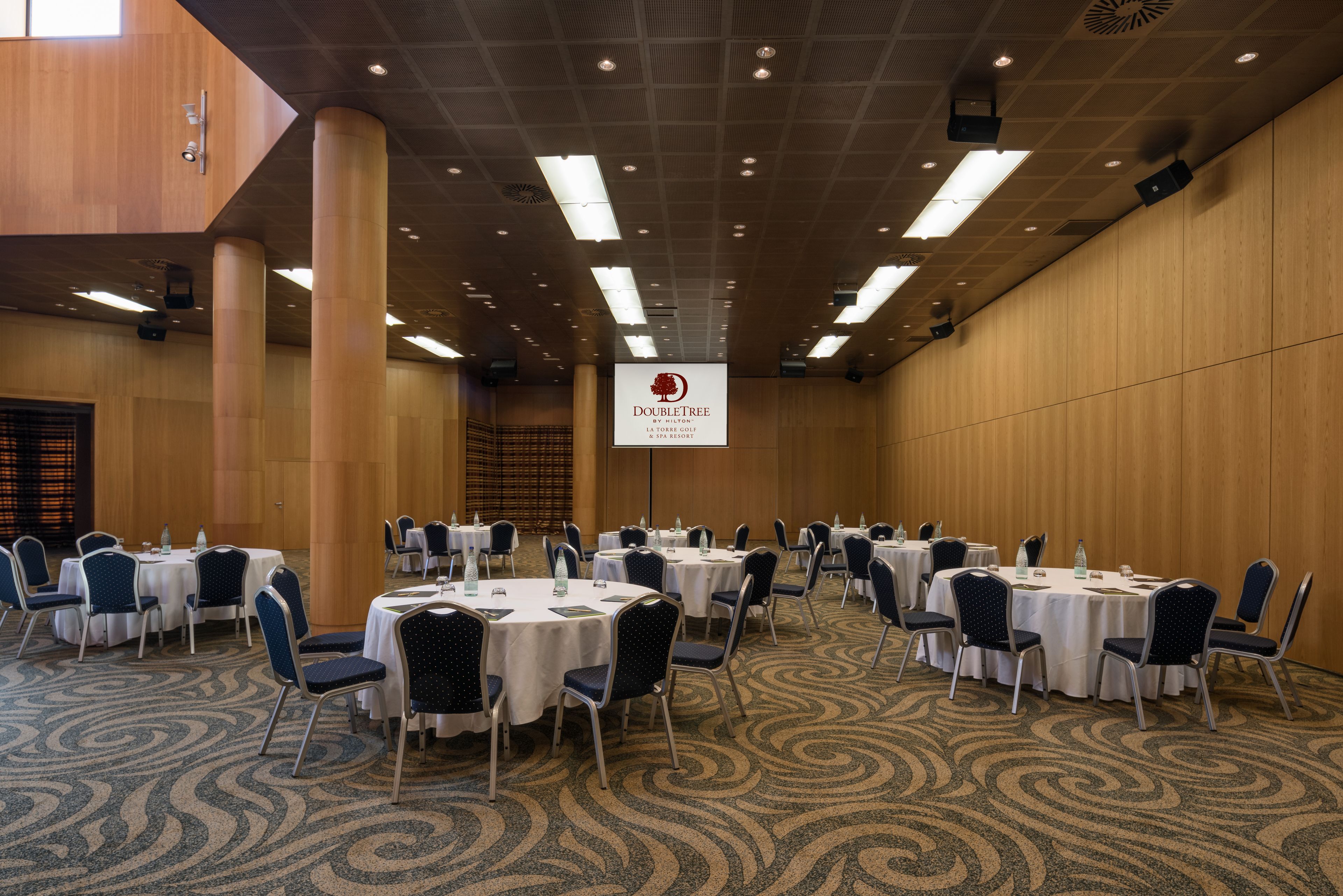 A conference room with round tables and chairs, a wooden wall, and a ceiling with recessed lighting.