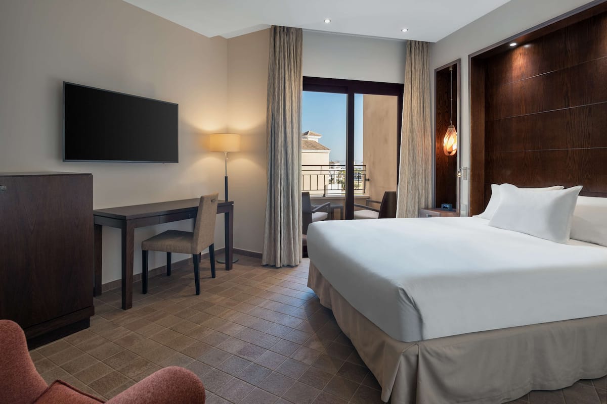 A hotel room with a bed, desk, chair, TV, and a view of the outside.