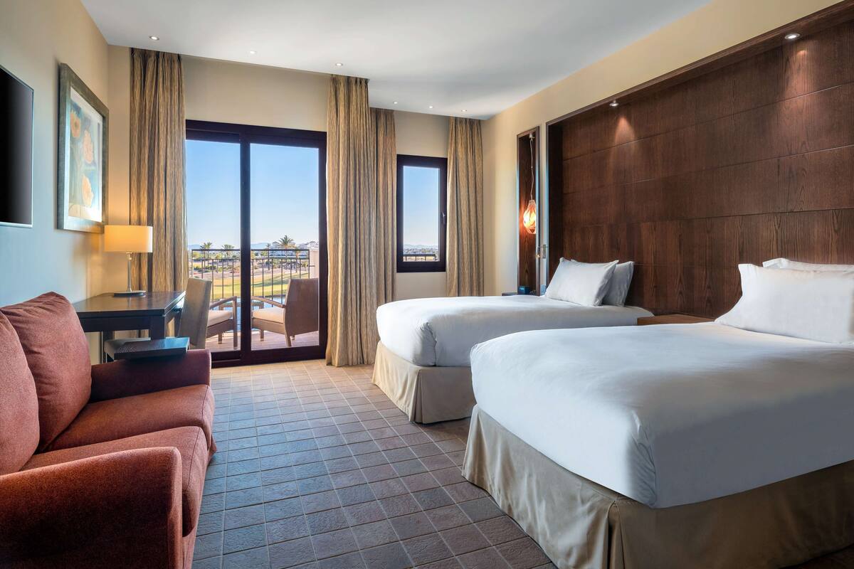 A hotel room with two beds, a sofa, a desk, and a view of the outdoors.