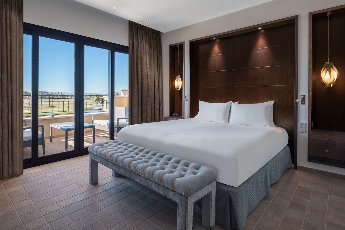 A large bed with a bench, wooden headboard, and a view of a terrace with outdoor furniture.