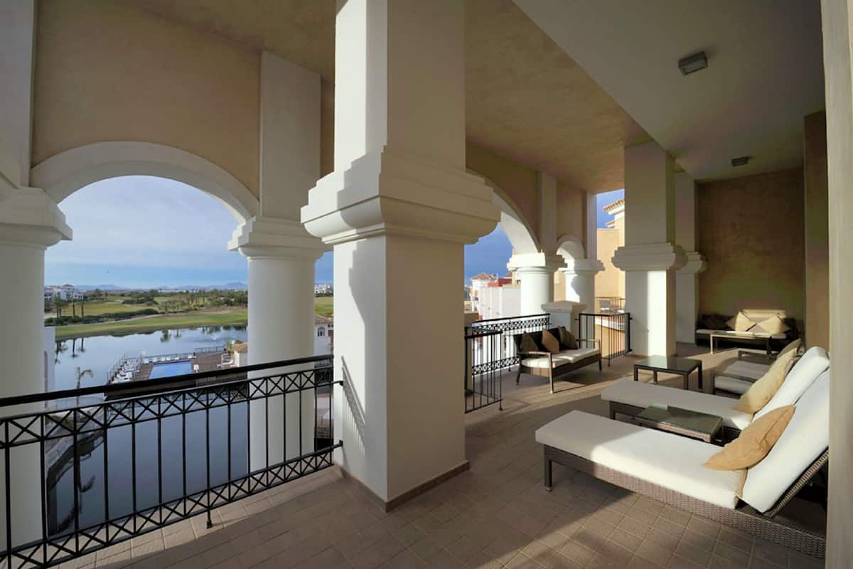 A balcony with a view of a lake and buildings, furnished with a sofa and chairs.