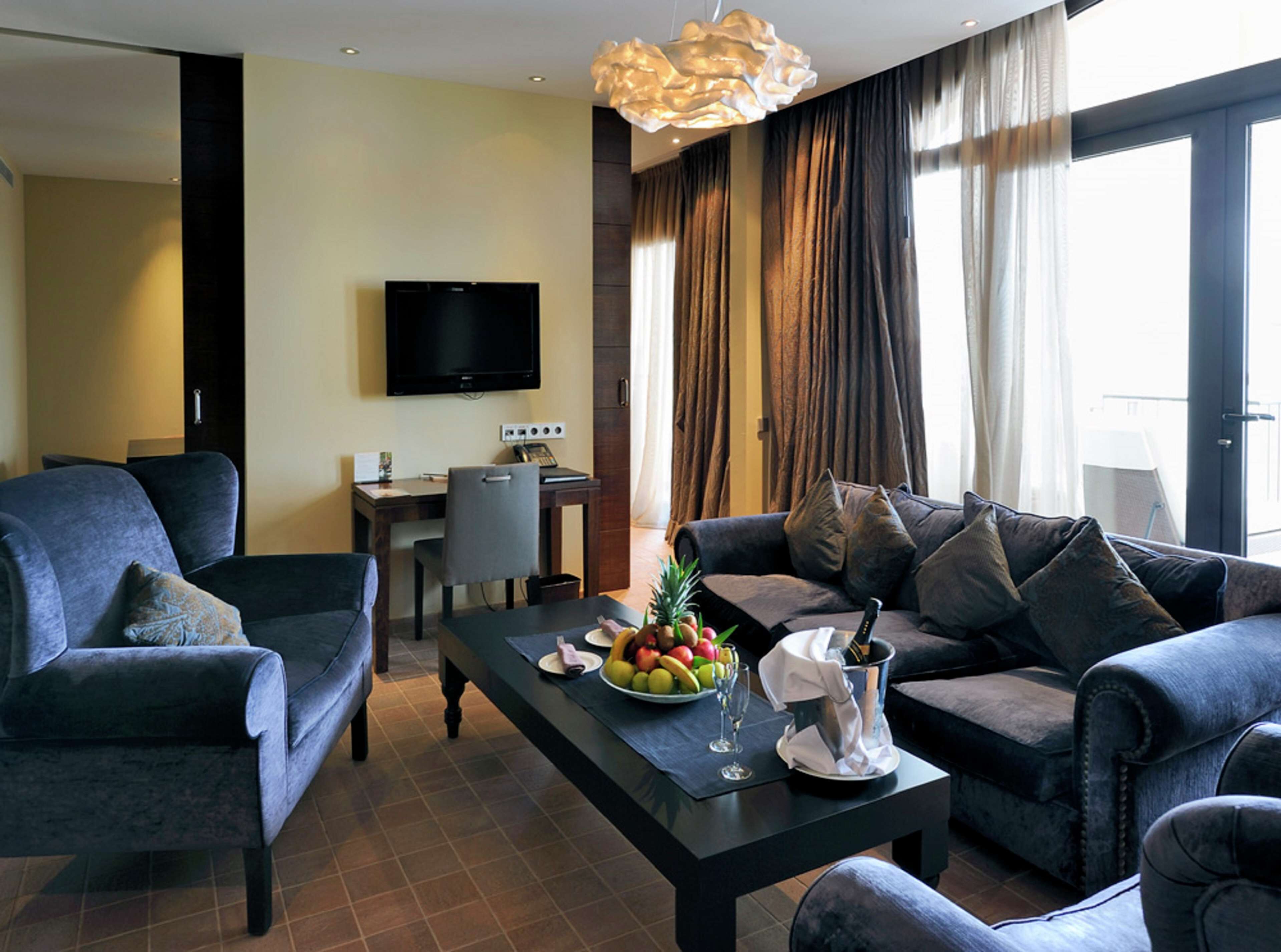A modern hotel room with a sofa set, a dining table with fruit and a bottle, a TV, and a desk with a chair.