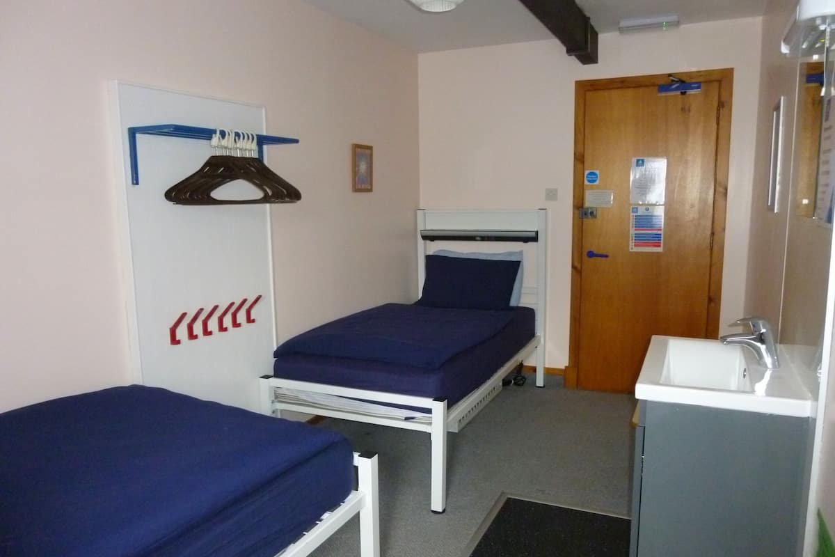 A small, compact room with two single beds, a wooden door, a white sink, and a mirror.