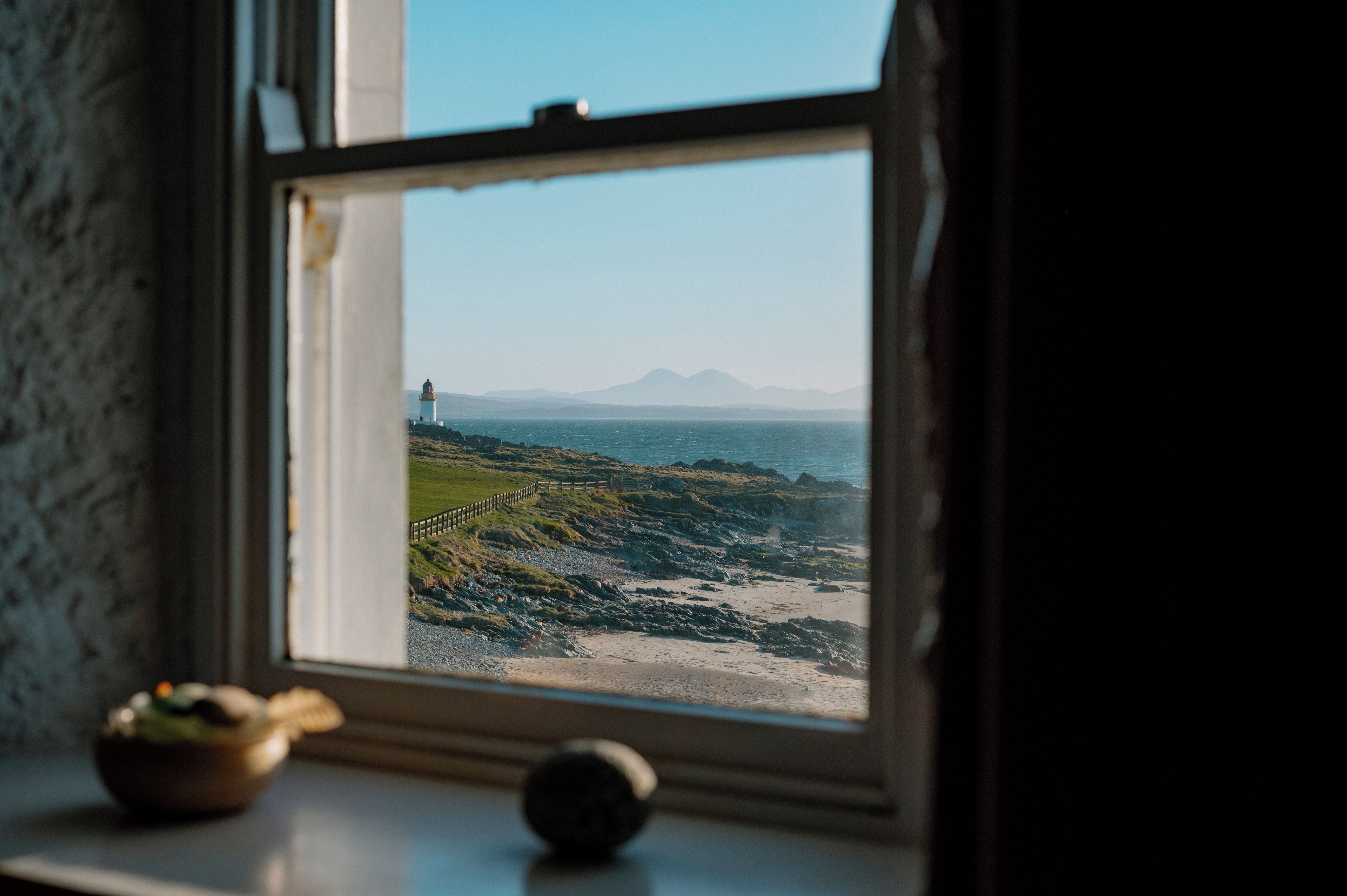 A coastal view through a window with a lighthouse in the distance, a rocky shoreline, and a grassy area.