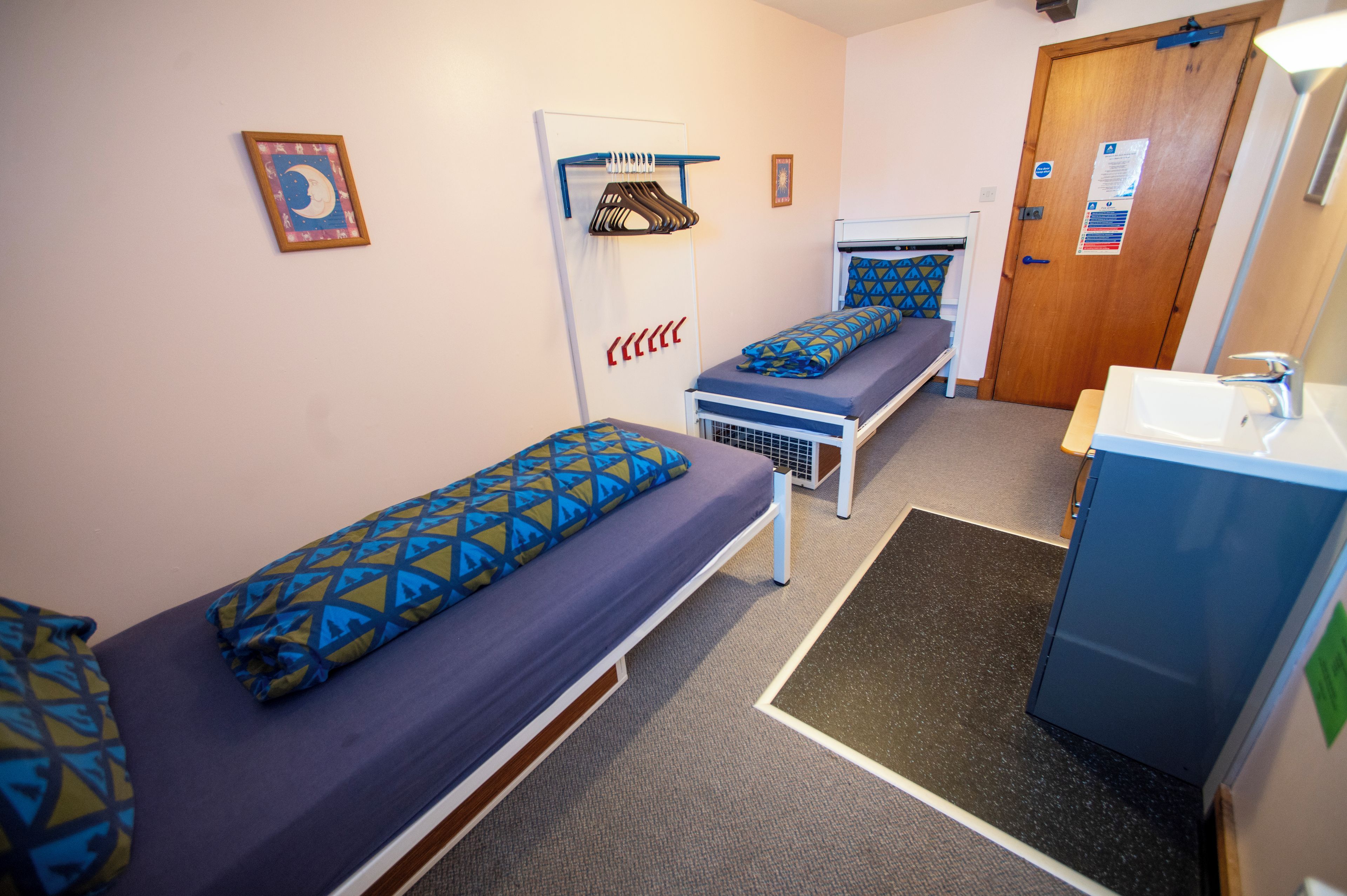 Habitación tipo dormitorio con dos camas individuales, una mesita pequeña, un banco, un lavabo y una puerta.