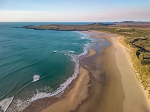 Vista aérea de un paisaje costero con una playa arenosa, aguas azul cristalino y colinas onduladas al fondo.