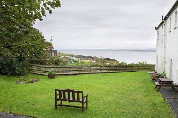A grassy outdoor area with a bench, a picnic table, and a view of the sea. - Port Charlotte Youth Hostel (Islay Island)