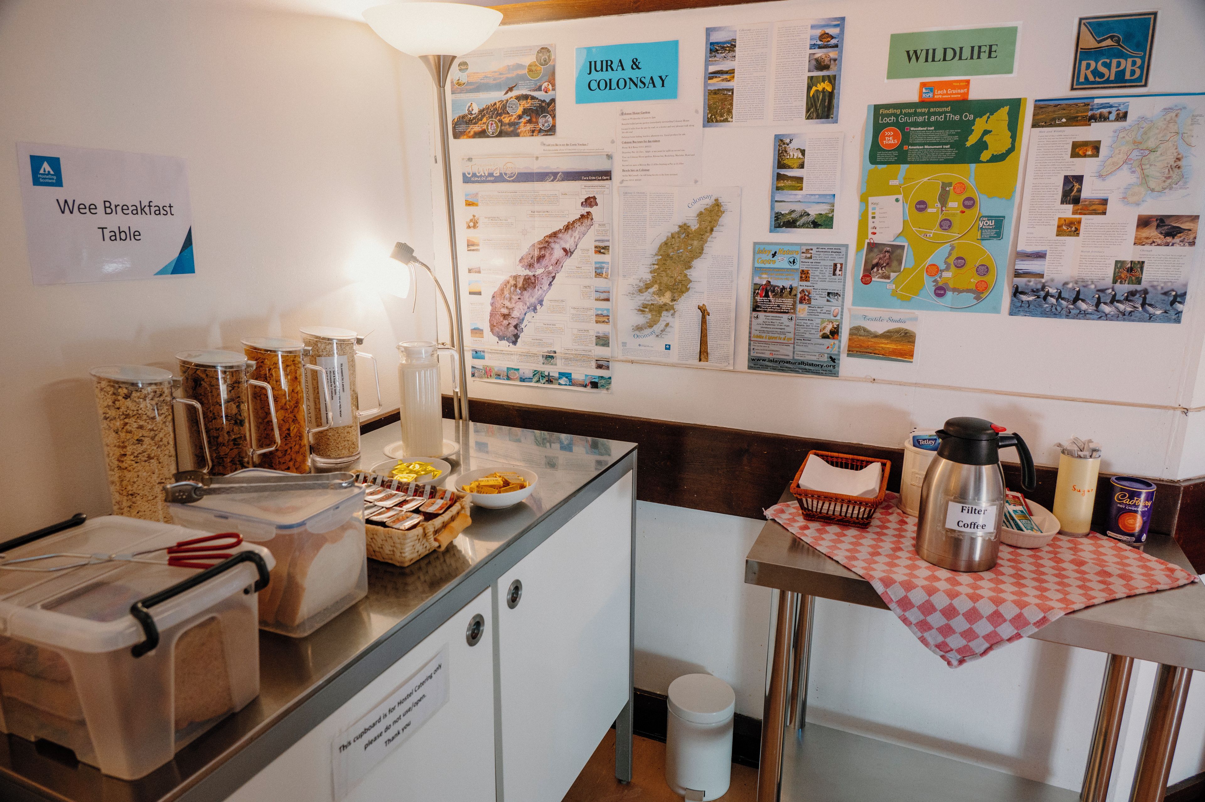 En frukosthörna med ett litet frukostbord, burkar med mat, en kaffekanna och informationsaffischer på väggen.
