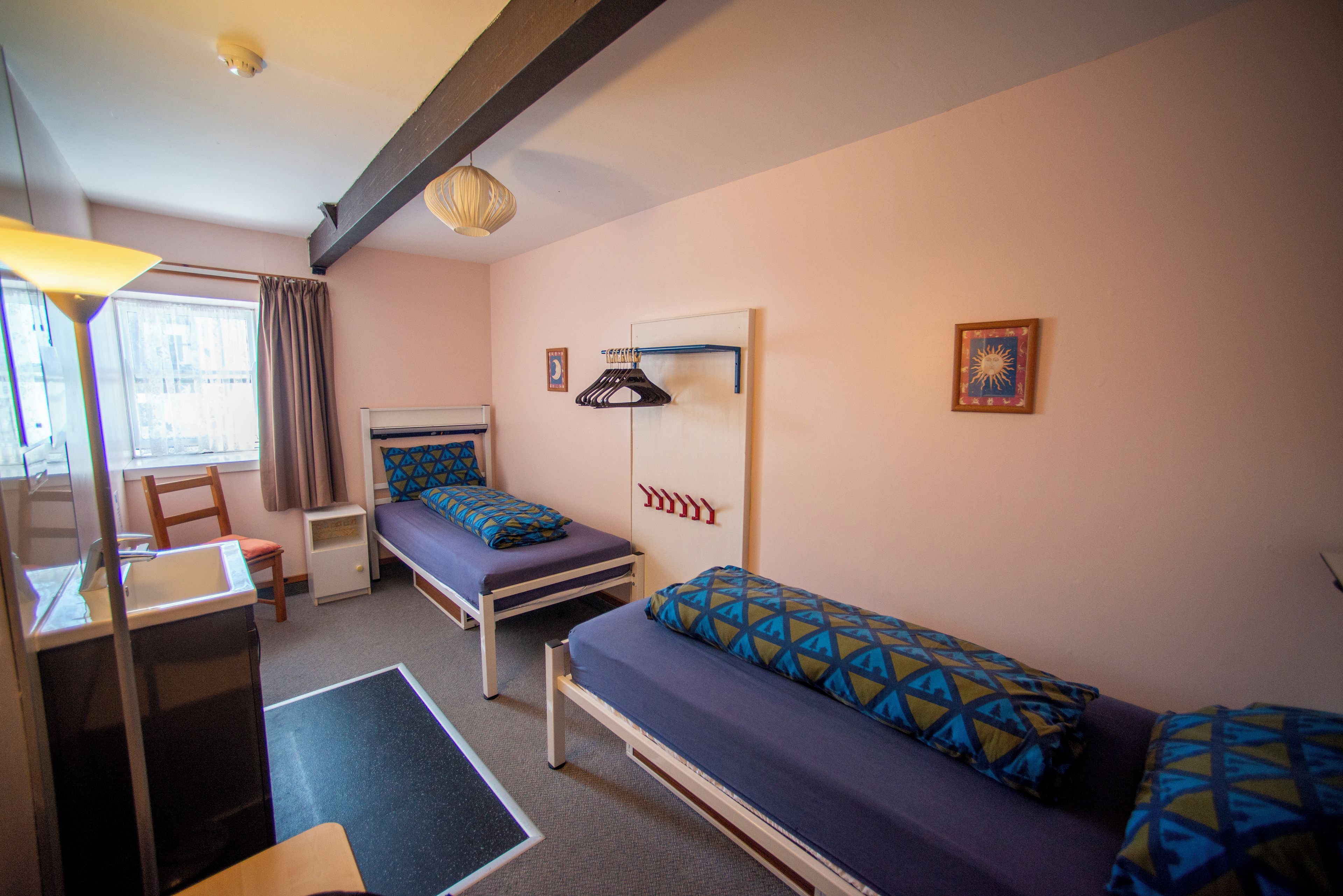 Une chambre de dortoir avec deux lits simples, un bureau avec une chaise, un luminaire suspendu et une fenêtre avec des rideaux.