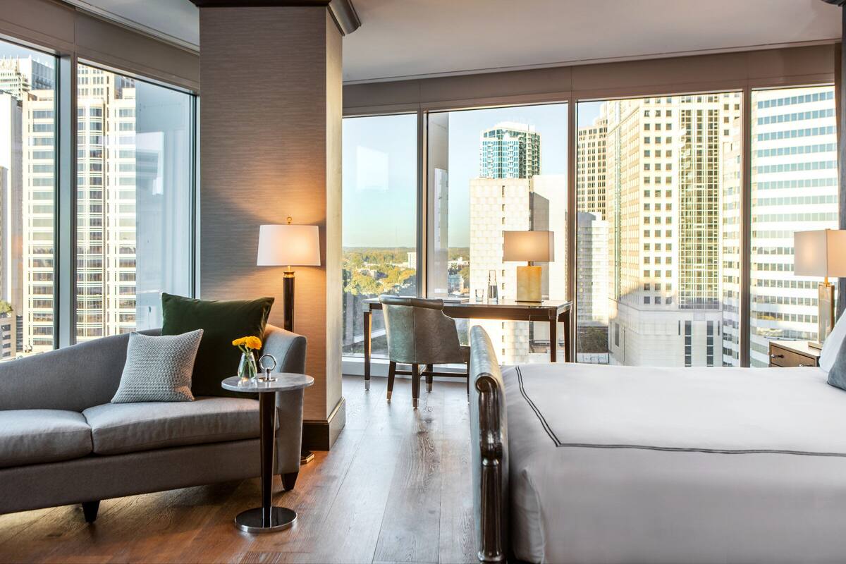 A hotel room with a large bed, a sofa, a small table with a lamp, and a chair. There are large windows offering a city view.