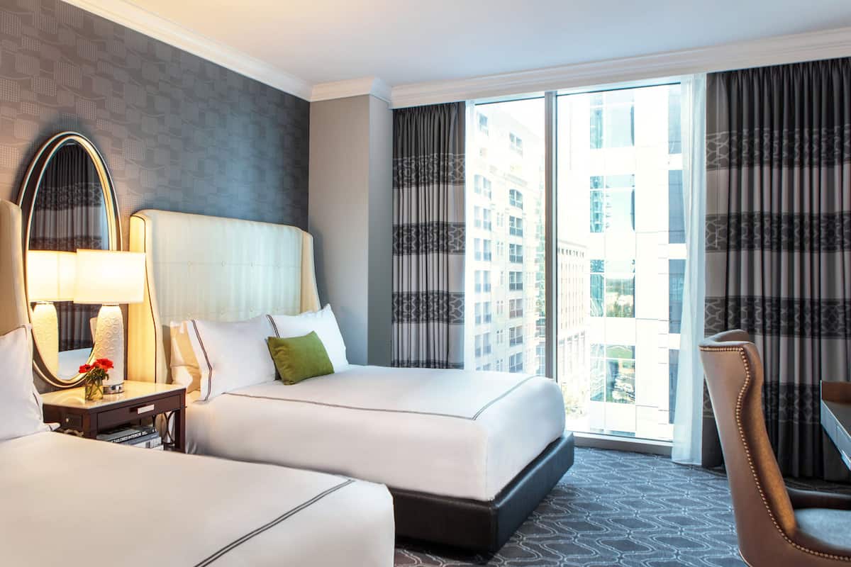 A hotel room with two beds, a large mirror, a bedside table with a lamp, and a view of the city through large windows.