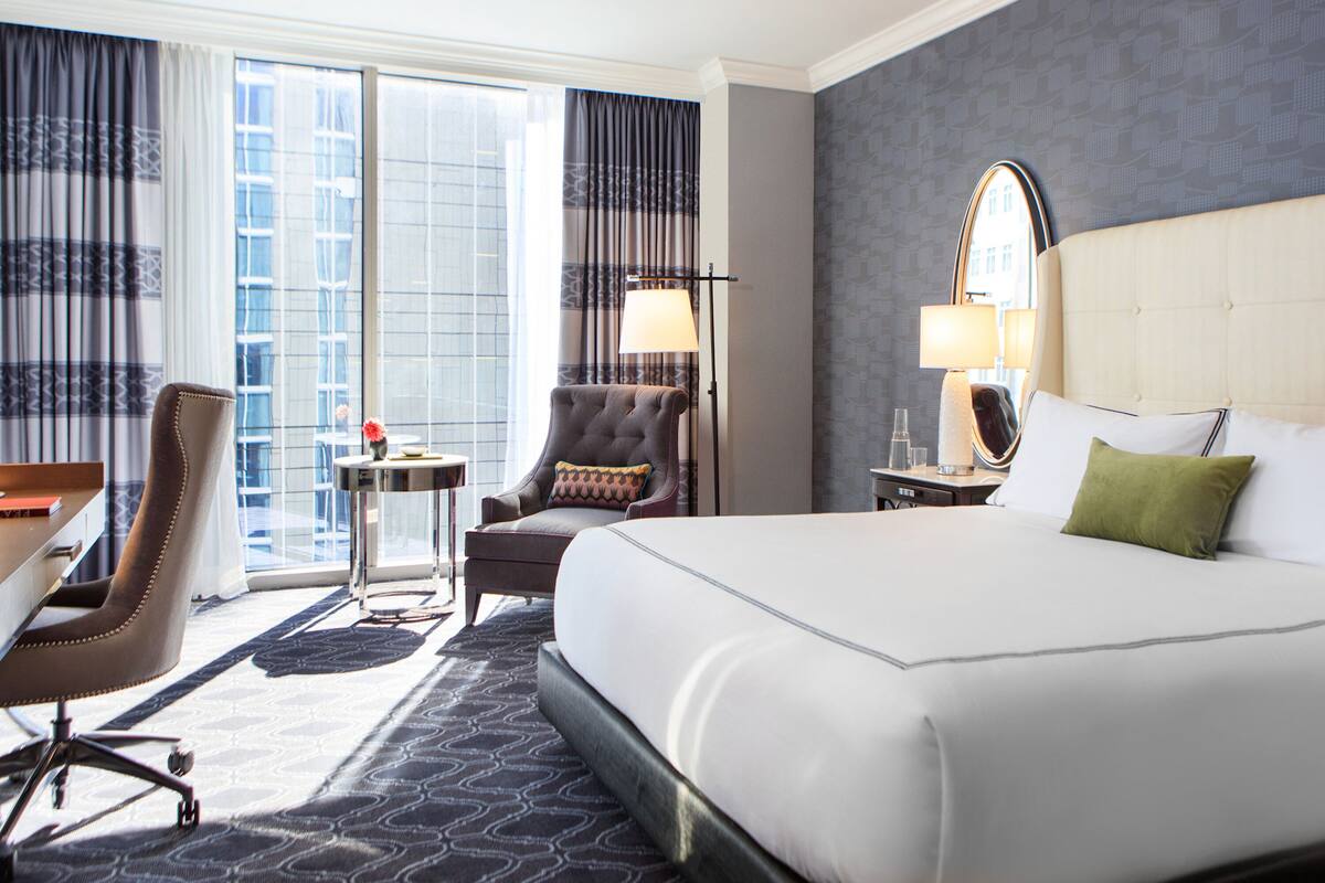 A hotel room with a large bed, a desk, a chair, a small table, and a window with curtains.