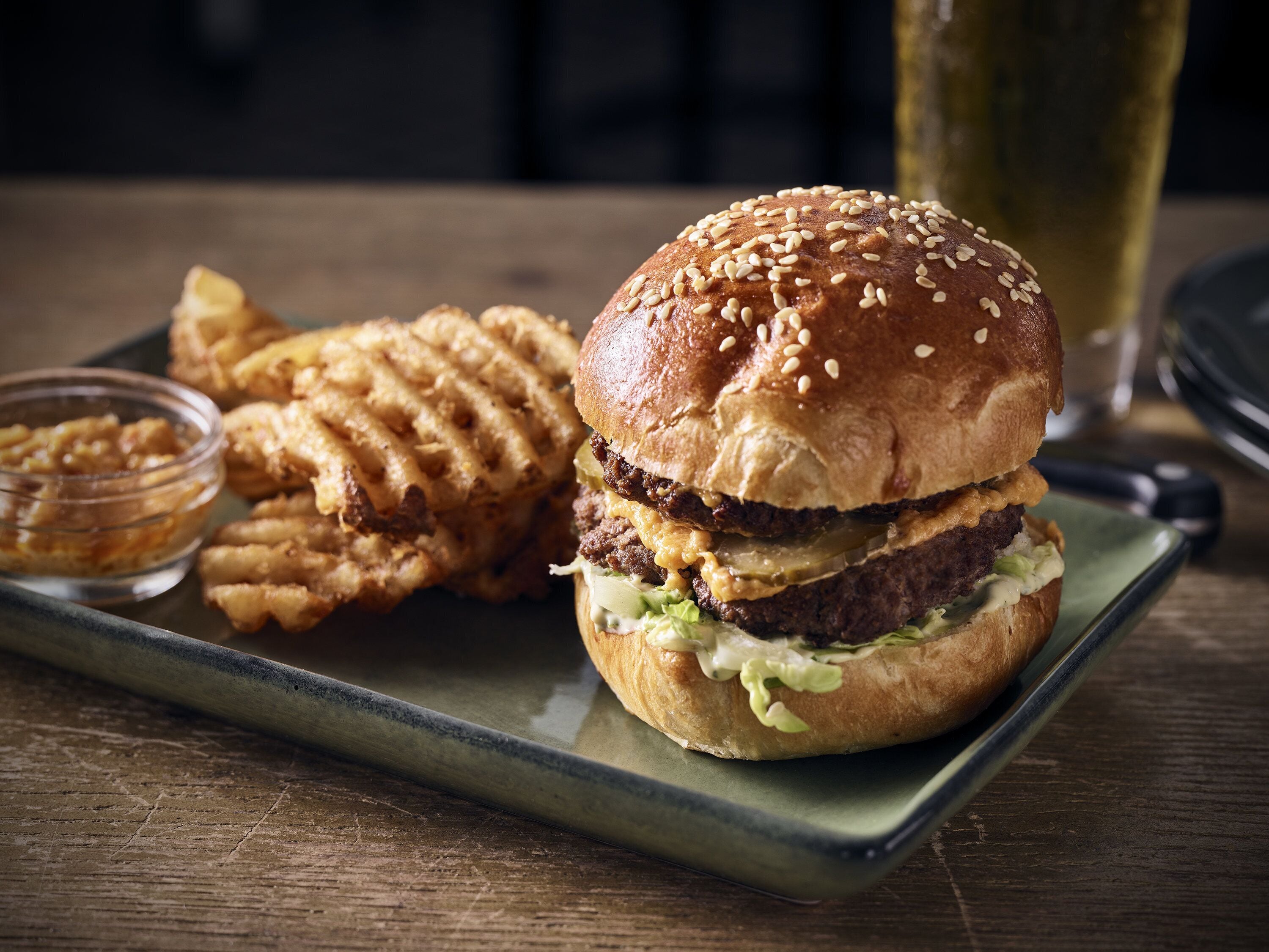 Un hamburger aux graines de sésame, servi dans un pain, accompagné de frites et d’un petit bol de sauce sur une assiette verte.