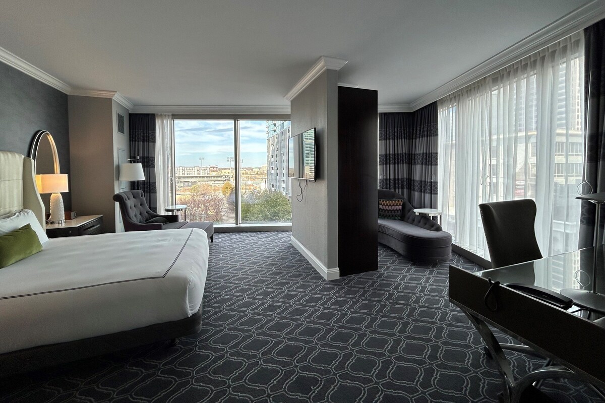 A hotel room with a large bed, a desk, a chair, a sofa, and a view of the city.
