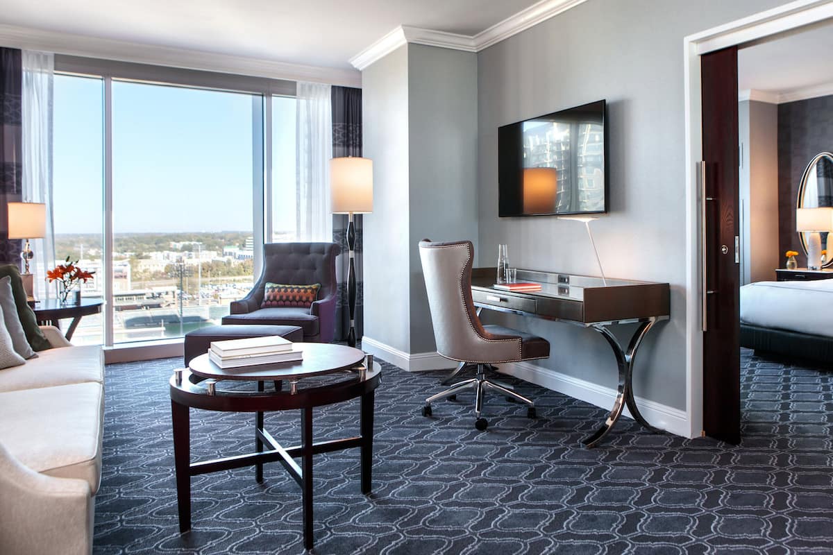 A hotel room with a large window, a desk, a chair, a sofa, and a TV.
