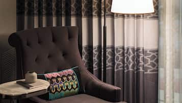 Quarto de hotel com poltrona escura, mesa de apoio com um livro e um vaso, e luminária de chão.